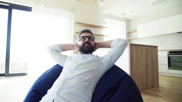 Cheerful mature man jsitting on bean bag in unfurnished house, moving in new home concept. Slow motion.