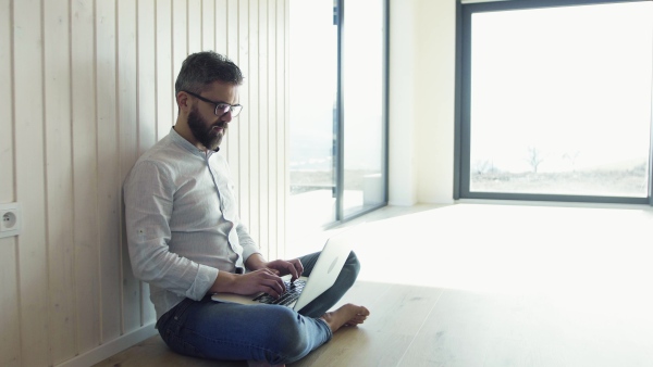 A mature man sitting on the floor in unfurnished new house, using laptop. A new home concept. Copy space.