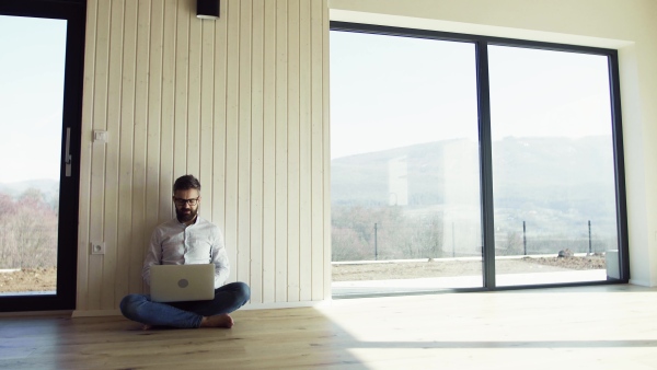 A mature man sitting on the floor in unfurnished new house, using laptop. A new home concept. Copy space.