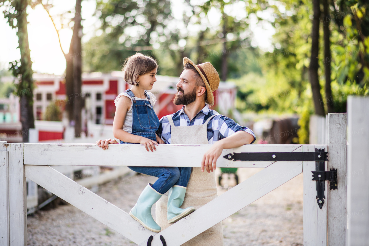 A portrait of father with small daughter outdoors on family farm, standing by wooden gate.