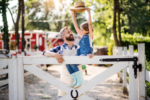 A portrait of father with small daughter outdoors on family farm, standing by wooden gate.