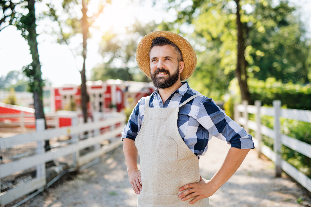 A portrait of mature man farmer with hat and apron standing outdoors on family farm.