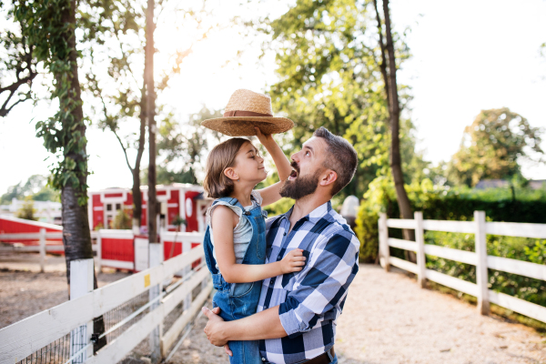 A happy father with small daughter standing outdoors on family farm, playing with hat.