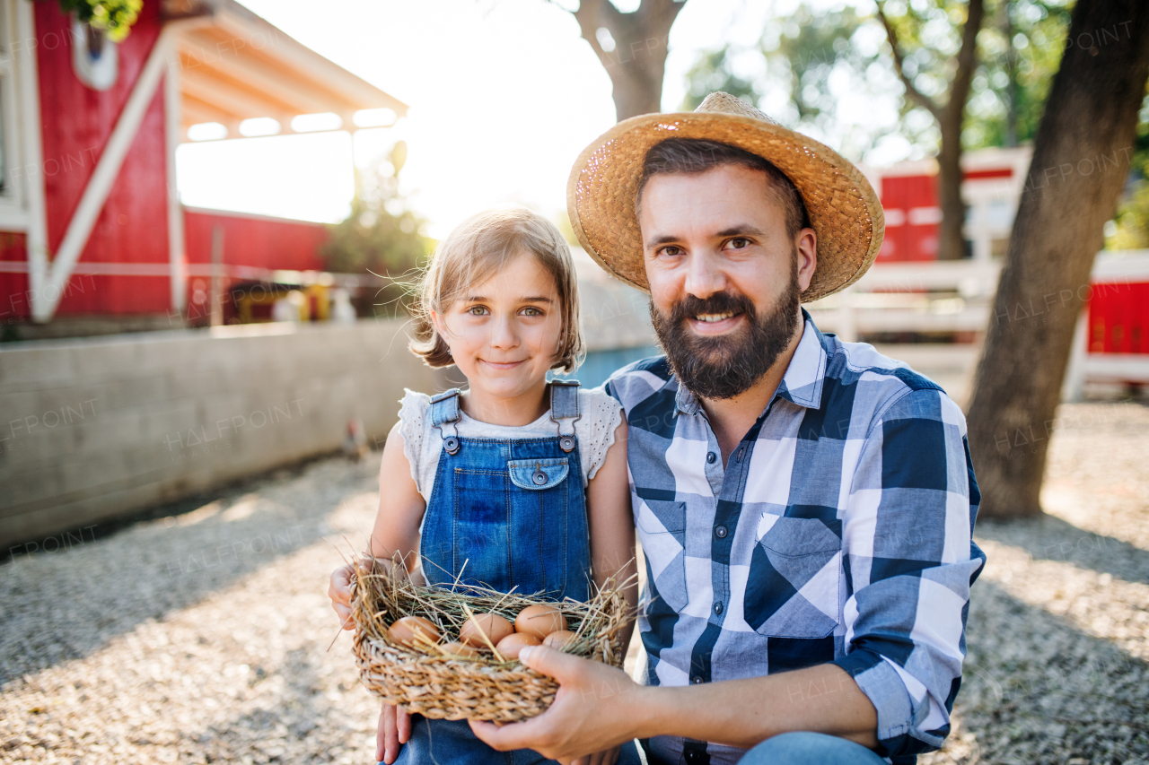 A father with small daughter outdoors on family farm, holding basket with eggs.