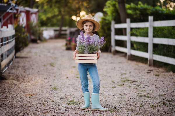 A front view of small girl with a hat standing outdoors on family farm, holding plants.