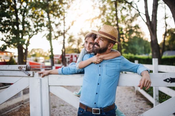 A portrait of father with small daughter outdoors on family farm, standing by wooden gate.