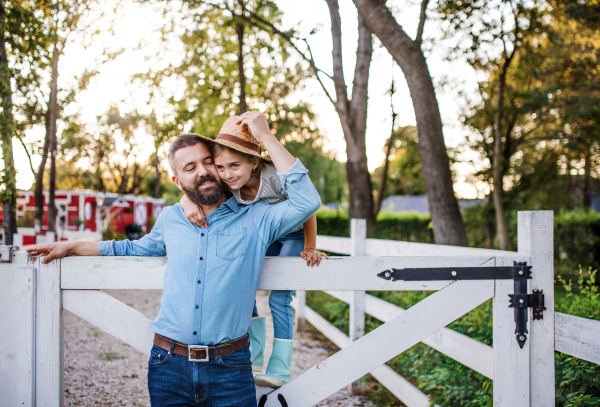 A portrait of father with small daughter outdoors on family farm, standing by wooden gate.