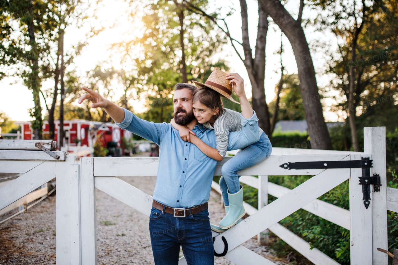 A portrait of father with small daughter outdoors on family farm, standing by wooden gate.