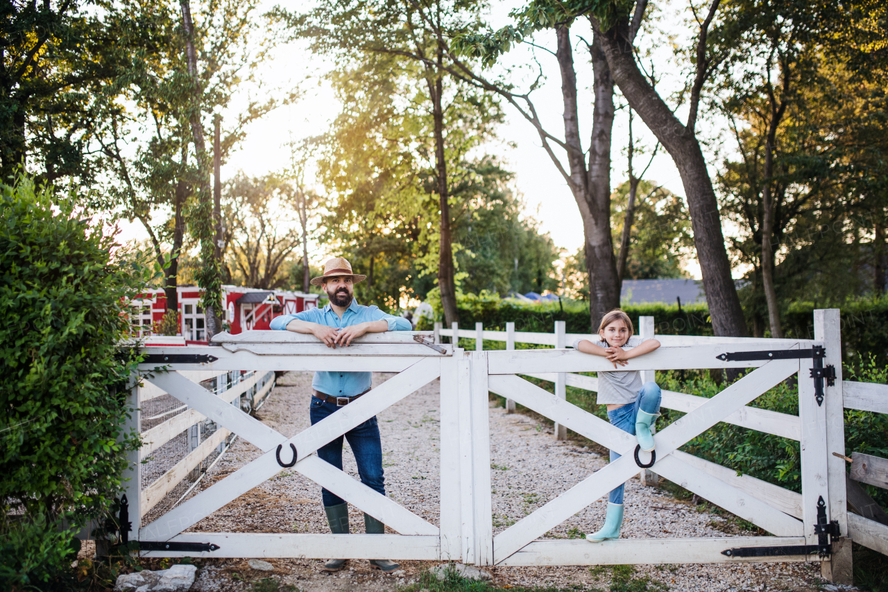 A portrait of father with small daughter outdoors on family farm, looking at camera.