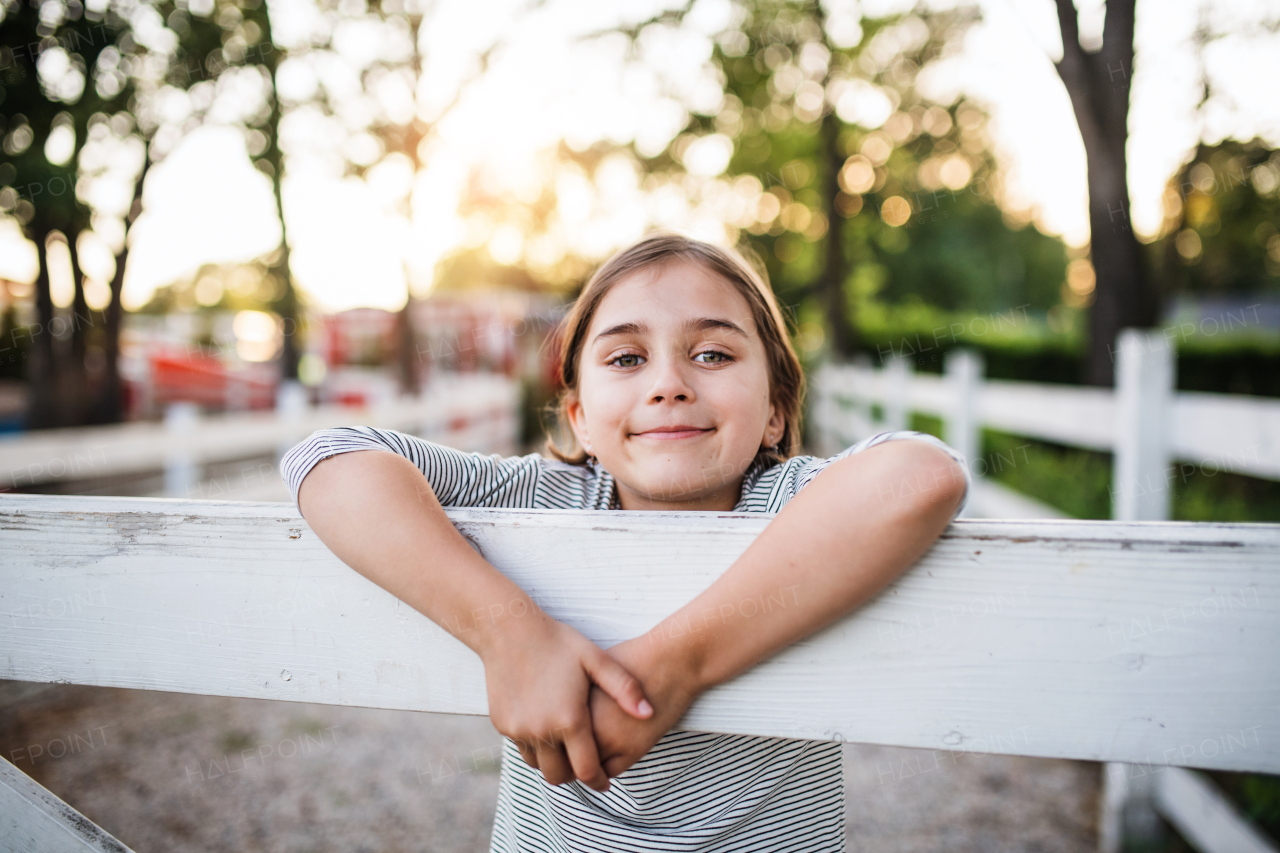 A portrait of happy small girl on family farm, standing by wooden gate.