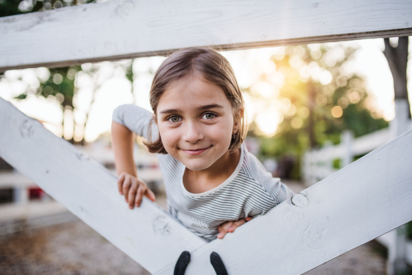 A portrait of happy small girl on family farm, standing by wooden gate.