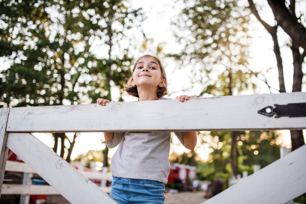 A portrait of happy small girl on family farm, standing by wooden gate.