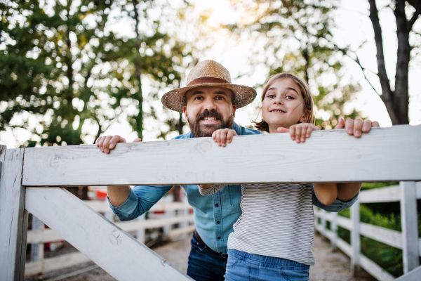 A portrait of father with small daughter outdoors on family farm, looking at camera.