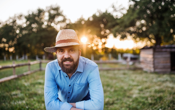 A portrait of happy mature man farmer with hat and apron standing outdoors on family farm at sunset.