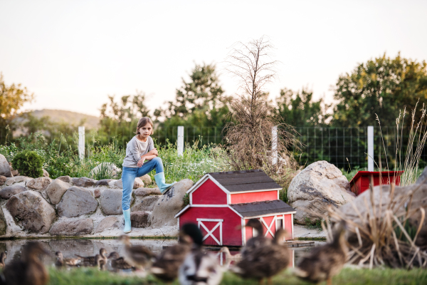 A front view of small girl with a hat standing outdoors on family farm, resting.