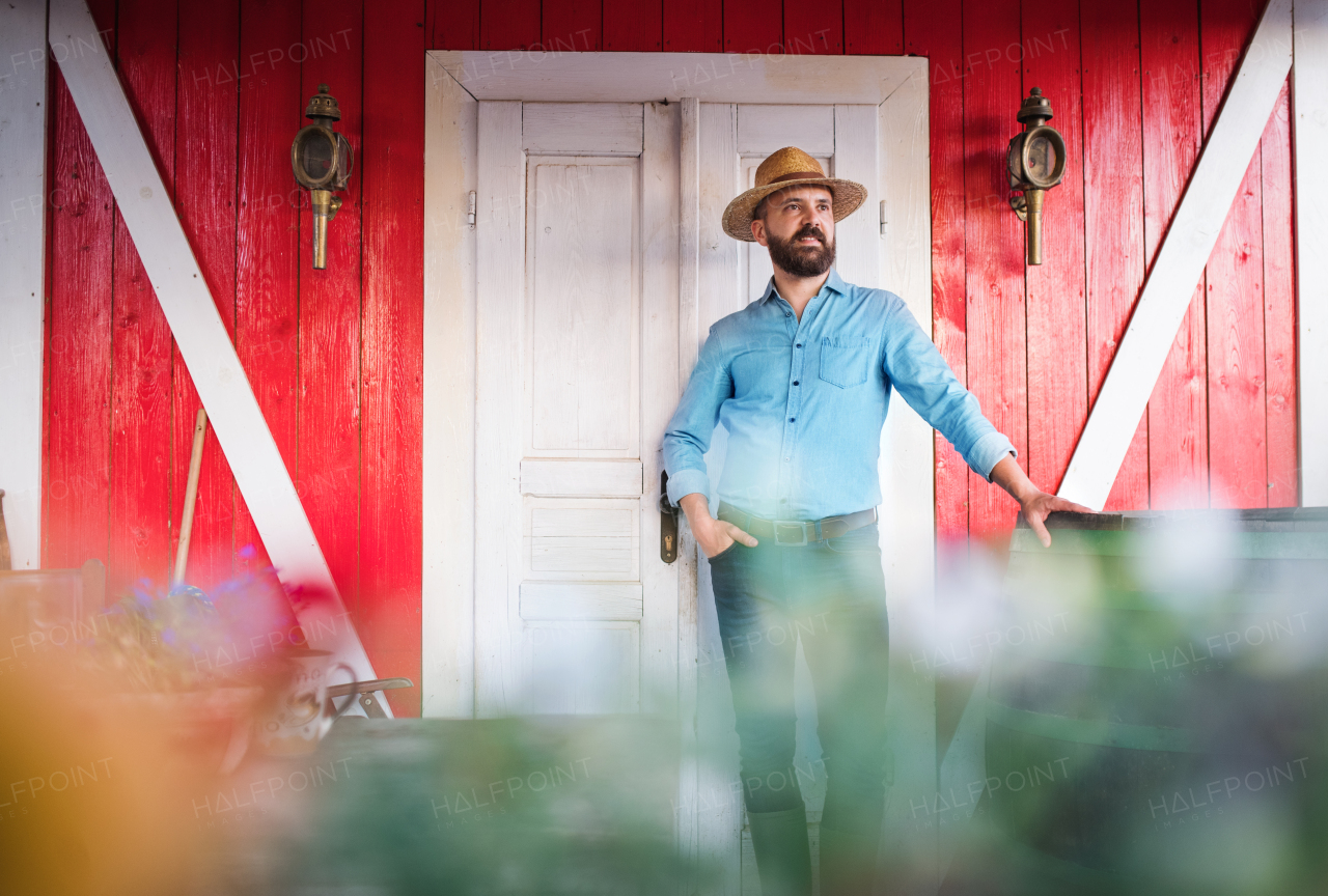 A portrait of mature man farmer with a hat standing outdoors in front of family farm.