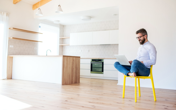 A mature man sitting on chair in unfurnished new house, using laptop. A new home concept.