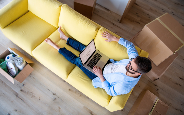 Top view of mature man sitting on sofa in unfurnished house, using laptop. A moving in new home concept.