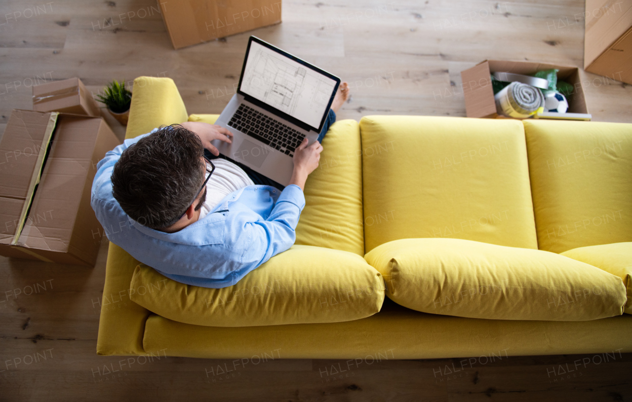 Top view of mature man sitting on sofa in unfurnished house, using laptop. A moving in new home concept.