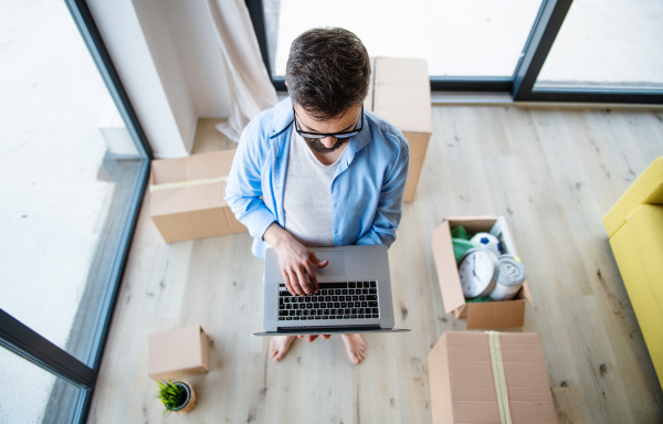 Top view of mature man with boxes moving in new house, sitting and using laptop.