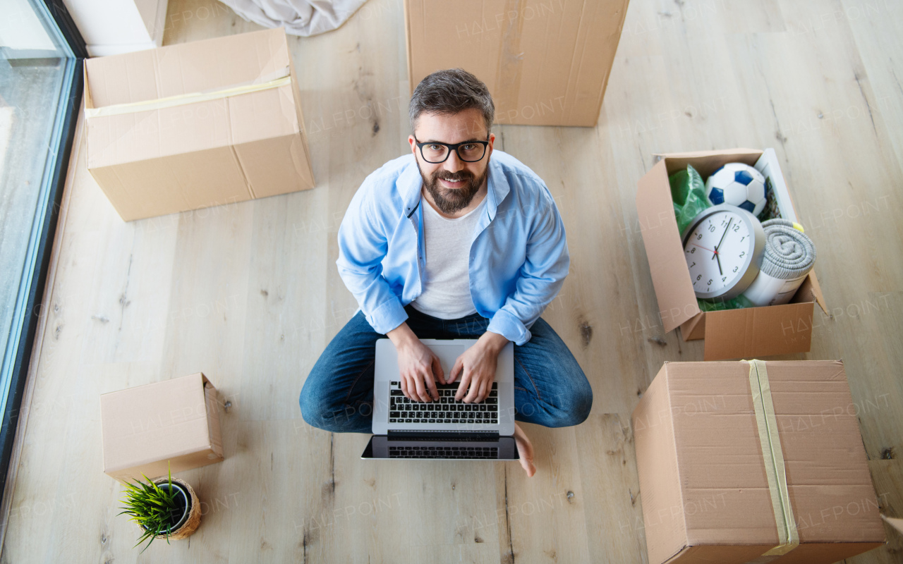 Top view of mature man with boxes moving in new house, sitting and using laptop.