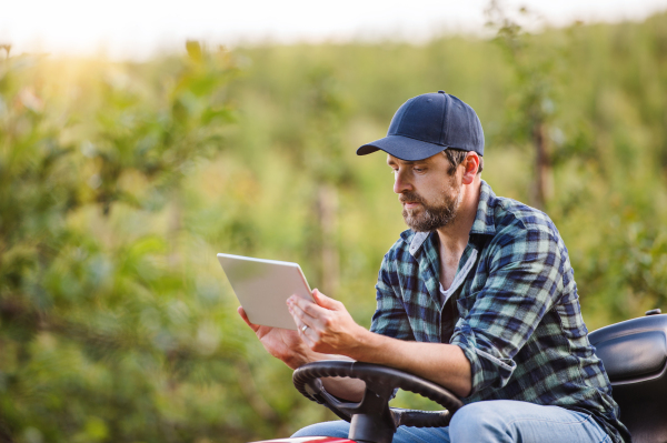 A serious mature farmer with tablet sitting on mini tractor outdoors in orchard.