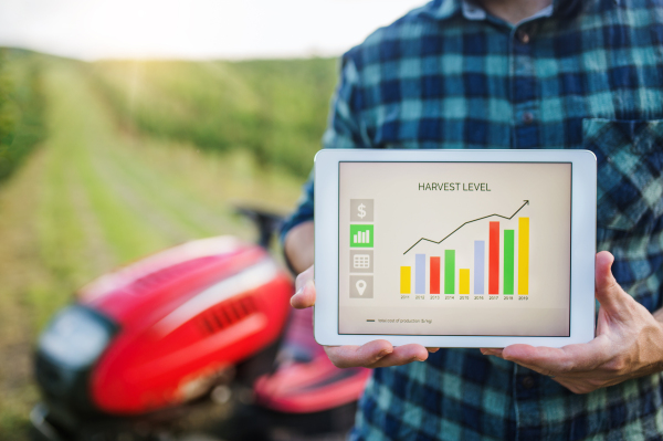 A midsection of farmer with tablet standing by mini tractor outdoors in orchard, checking harvest level.