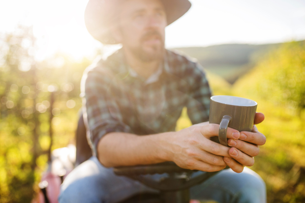 A front view of mature farmer with cup of coffee outdoors in orchard, resting.