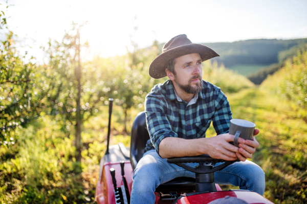 A front view of mature farmer with cup of coffee outdoors in orchard, resting.