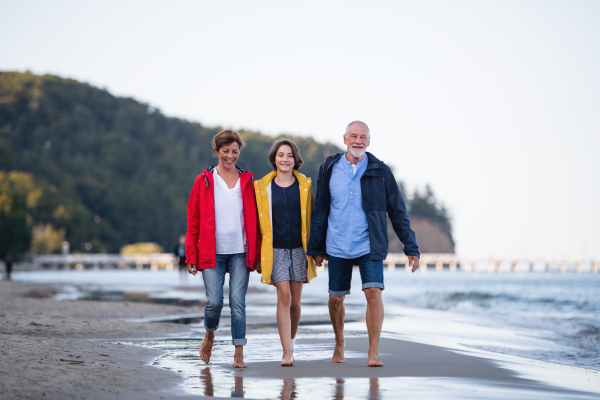 A senior couple holding hands with their preteen granddaughter and walking on sandy beach.