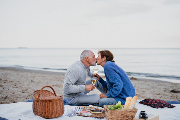 A happy senior couple in love sitting on blanket and kissing when having picnic outdoors on beach.