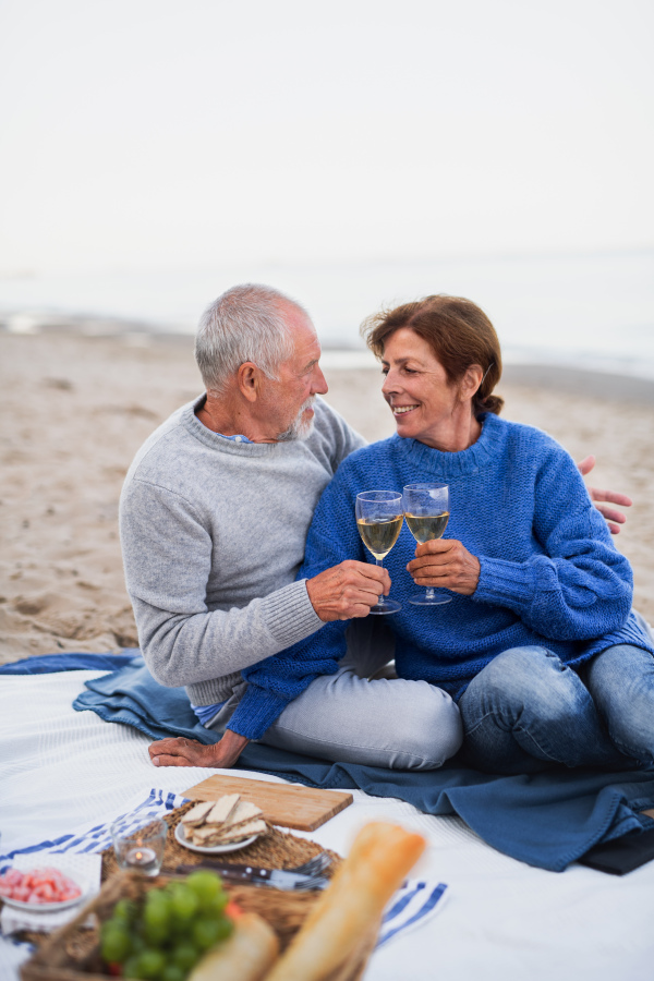 A happy senior couple in love sitting on blanket and having picnic outdoors on beach by sea.