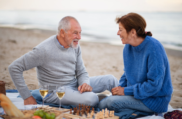 A happy senior couple in love sitting on blanket drinking wine and playing chess outdoors on beach.