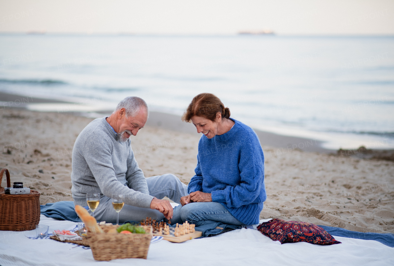 A happy senior couple in love sitting on blanket drinking wine and playing chess outdoors on beach.