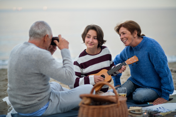 A happy senior couple with granddaughter sitting on blanket and having picnic outdoors on beach by sea.