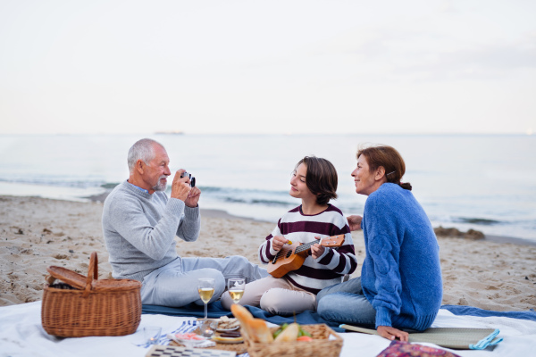 A happy senior couple with granddaughter sitting on blanket and having picnic outdoors on beach by sea.