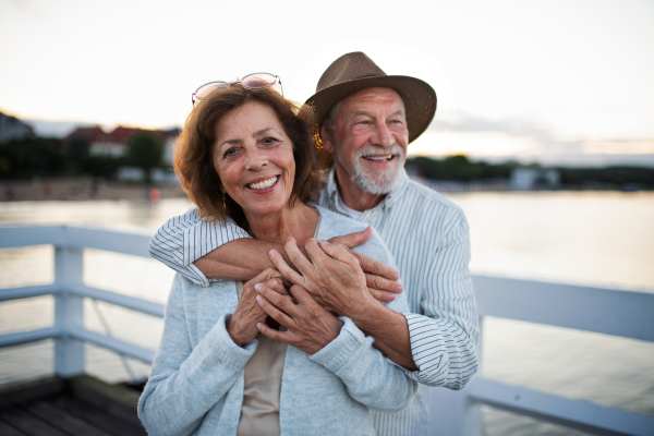 A happy senior couple hugging outdoors on pier by sea, looking at camera.