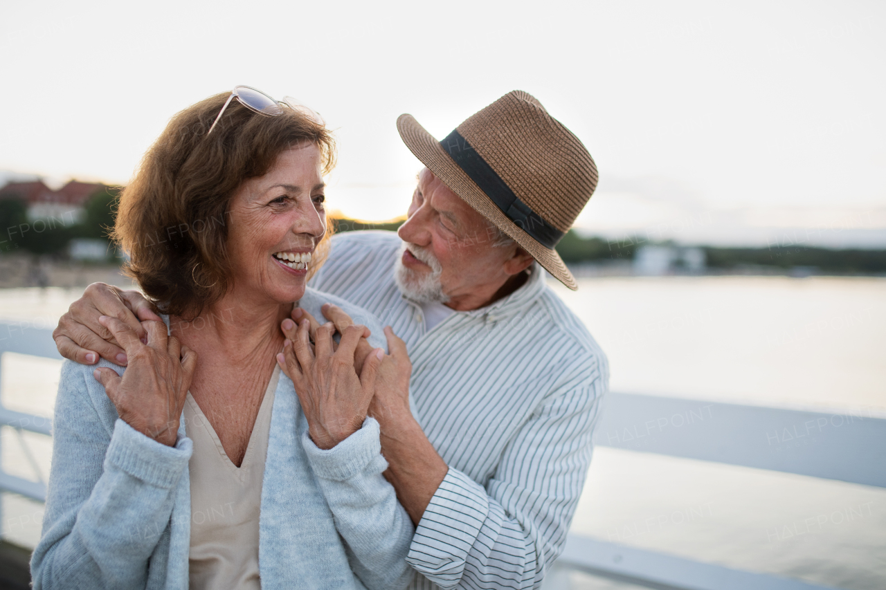 A happy senior couple hugging outdoors on pier by sea, looking at each other.