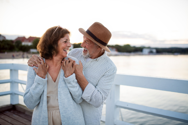 A happy senior couple hugging outdoors on pier by sea, looking at each other.