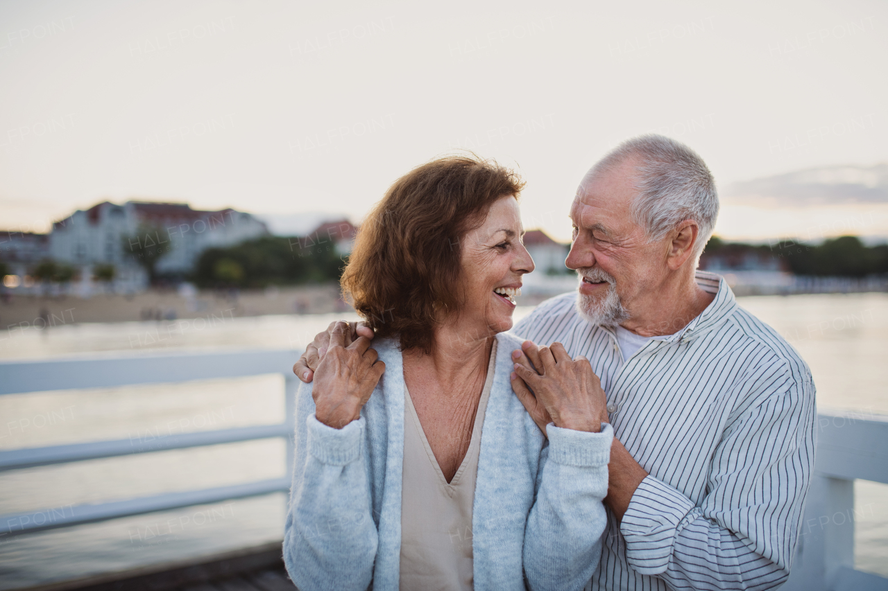 A happy senior couple hugging outdoors on pier by sea, looking at each other.