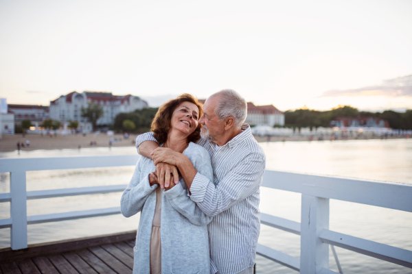 A happy senior couple hugging outdoors on pier by sea, looking at each other.