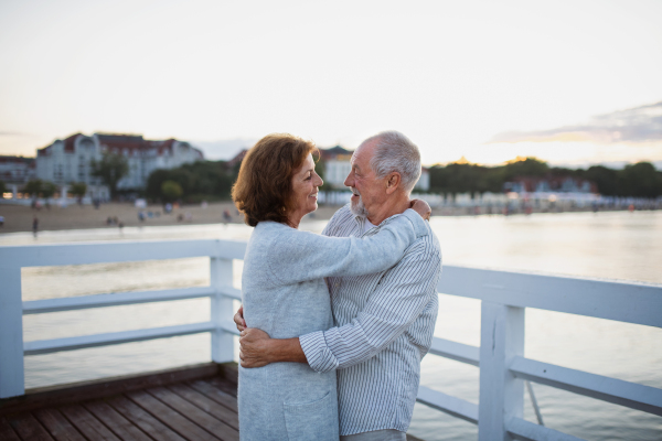 A happy senior couple hugging outdoors on pier by sea, looking at each other.