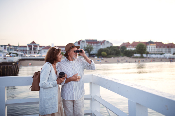 A happy senior couple hugging outdoors on pier by sea, looking at view through binoculars.