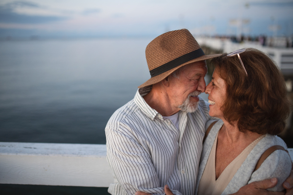 Portrait of happy senior couple in love hugging outdoors on a pier by sea, looking at each other, summer holiday.