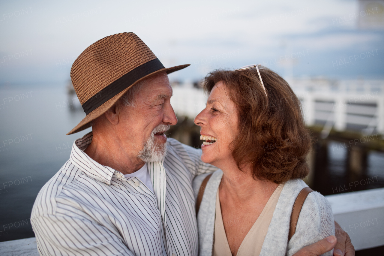 A happy senior couple hugging outdoors on pier by sea, looking at each other.