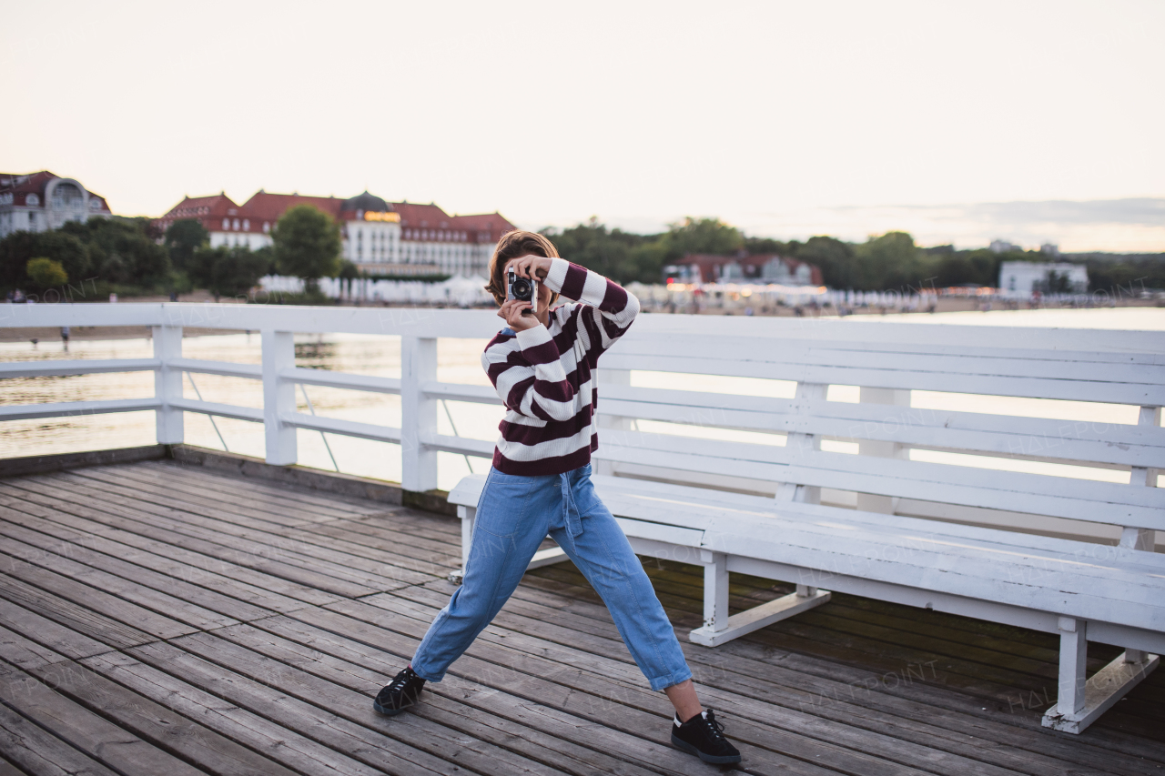 A portrait of preteen girl taking photo with camera on pier by sea at sunset, holiday concept.