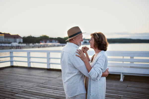 A happy senior couple dancing outdoors on pier by sea, looking at each other.