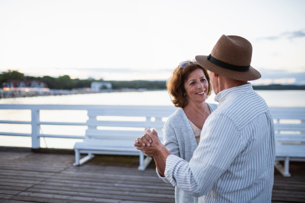 A happy senior couple dancing outdoors on pier by sea, looking at each other.