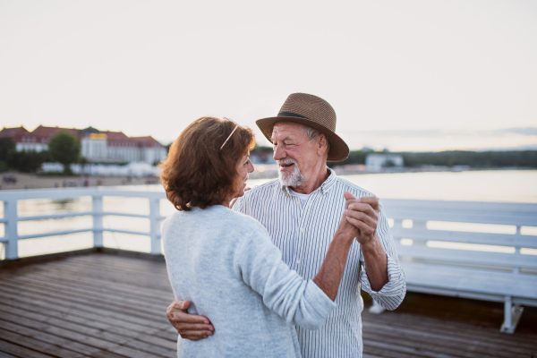 A happy senior couple dancing outdoors on pier by sea, looking at each other.