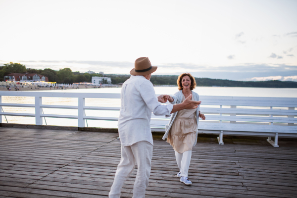 A happy senior couple dancing outdoors on pier by sea, looking at each other.